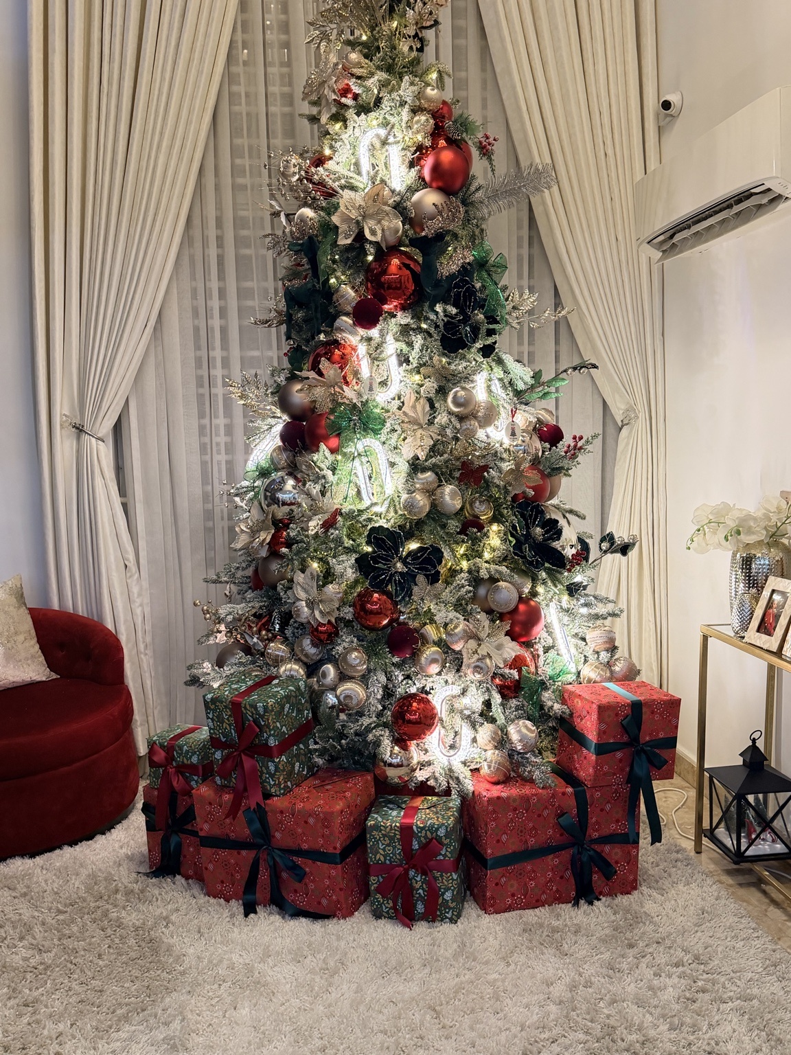 A flocked Christmas tree styled in champagne, red and velvet blooms with wrapped gifts arranged beneath it in a Lagos living room.