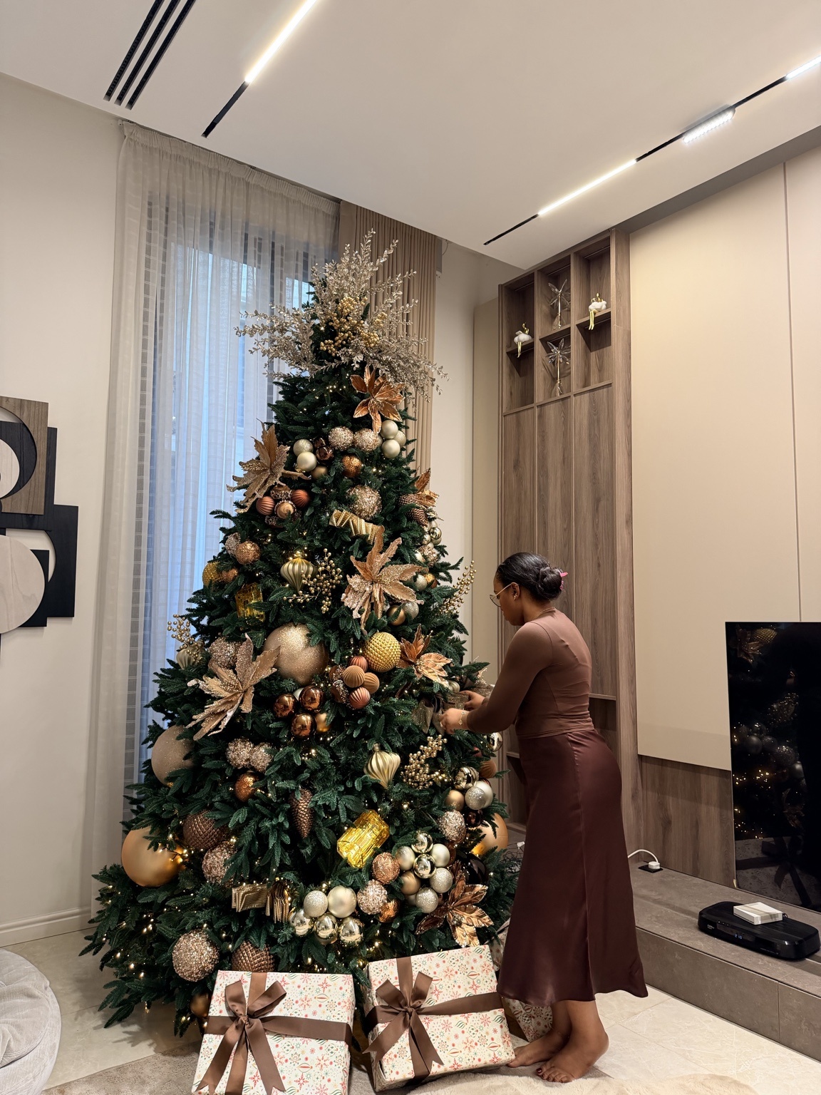 A stylist placing copper and gold ornaments on a full green Christmas tree in a warmly lit contemporary living room.